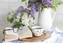 Table with book, eyeglasses, coffe cup and white and lavender flowers in a vase
