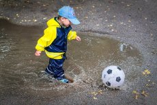 little boy in yellow and blue rain jacket playing with a ball in a water puddle or near a water's edge