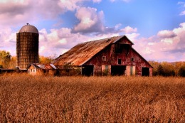 Rust-colored field in front of rust-colored barn and silo with blue sky and pinkish clouds