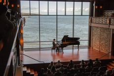 Pianist performing in front of an audience on a stage with a shiny wood floor and a background of blue water behind him 