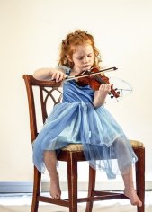 Red-haired, barefoot little girl in a blue dress with a serious expression sitting on a chair and playing a violin 