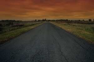 Dark grey road bordered by dark green fields leading to a distant point with a dark orange sky