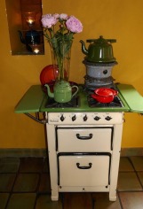An old-fashioned white oven with a green teapot, red crockery, and a vase of pink flowers on top